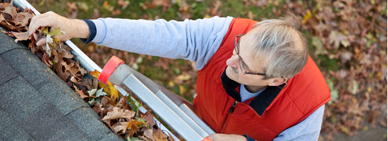An older man wearing glasses and a red vest stands on a ladder, clearing fallen leaves from a house gutter.