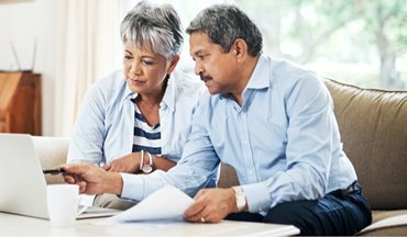 Two adults sit on a couch, looking at a laptop screen together. One points at the screen while the other holds papers.