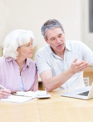 Two older adults sit at a table, looking at a laptop. The man gestures toward the screen while the woman takes notes in a notebook.