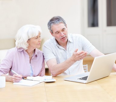 An older couple sits at a table, looking at a laptop. The man gestures toward the screen while the woman holds a pen and notebook, appearing to take notes.