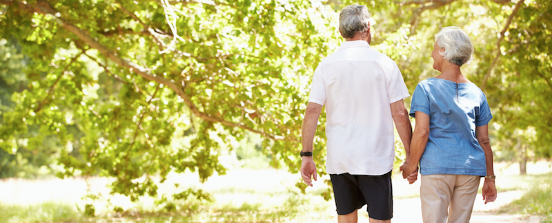 Two older adults walk hand in hand down a tree-lined path on a sunny day, surrounded by green foliage.