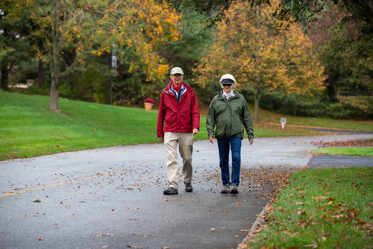 Two people in jackets and hats walk side by side on a paved path in a park surrounded by green grass and autumn trees.