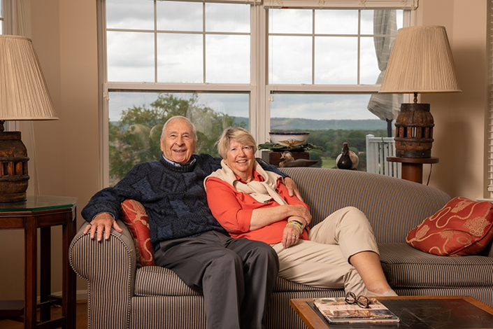An older man and woman sit closely together on a striped couch in a living room, smiling at the camera. Large windows and lamps frame the scene, showing a view of trees outside.