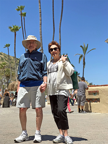 An older man and woman stand holding hands outdoors on a sunny day, with palm trees and people in the background.