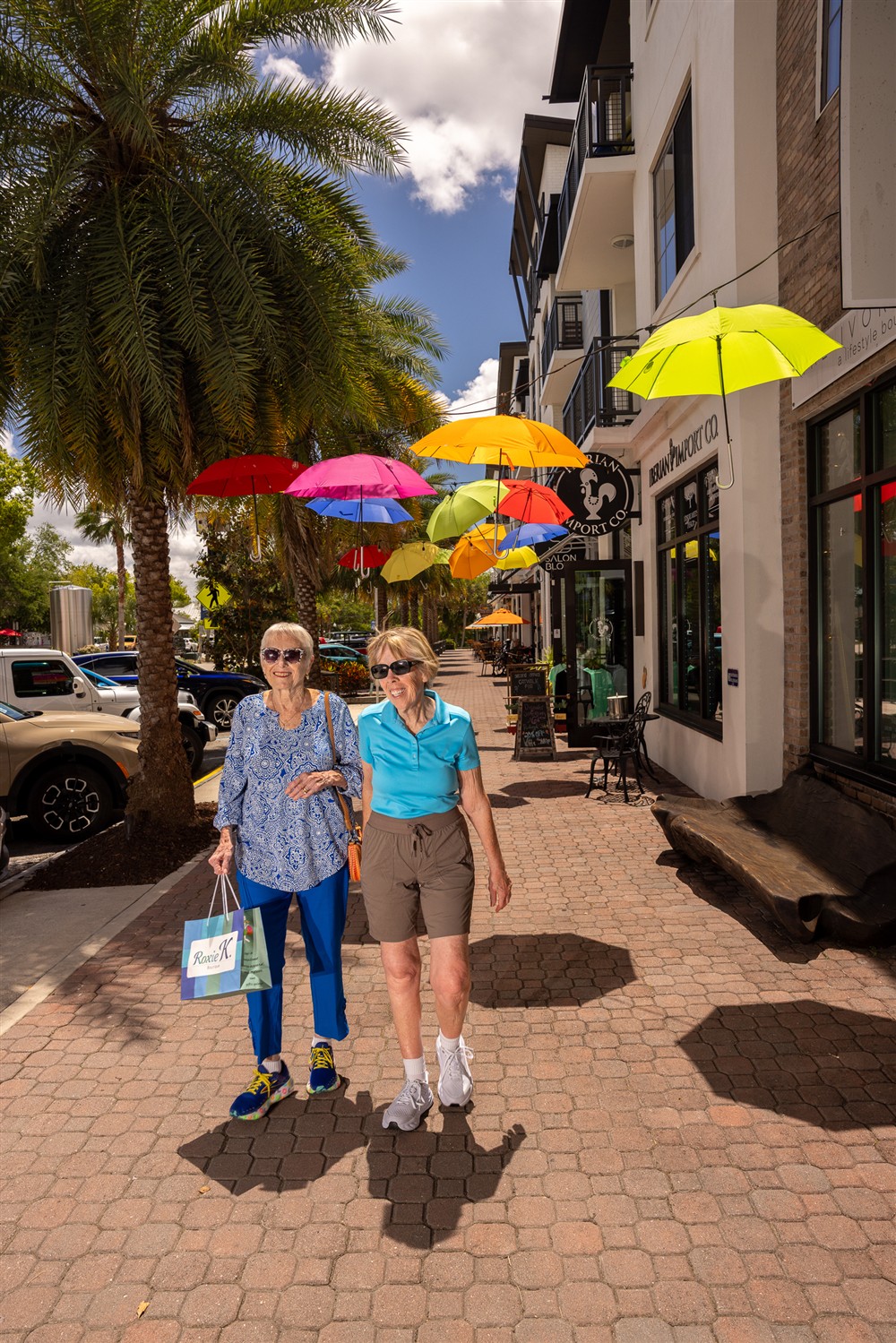 Two women walk on a sunny sidewalk beneath colorful hanging umbrellas, passing a café and palm tree, with one woman holding a shopping bag.