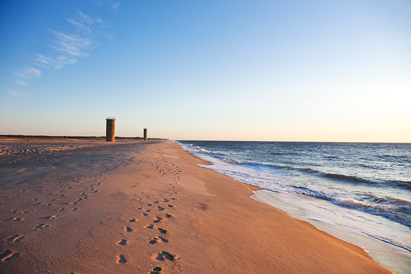 Sandy beach with footprints, calm ocean waves, and two cylindrical towers in the distance under a clear sky.