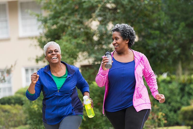 Two women in athletic wear walk and smile outdoors, one holding a water bottle. Green trees and a building are in the background.