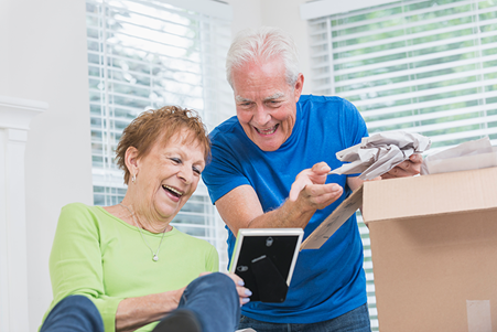 An older couple sits together, smiling and laughing while unpacking items from a cardboard box in a bright room with large windows.