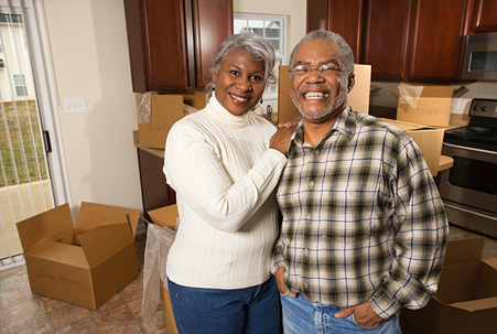 An older couple stands smiling in a kitchen surrounded by unpacked moving boxes.