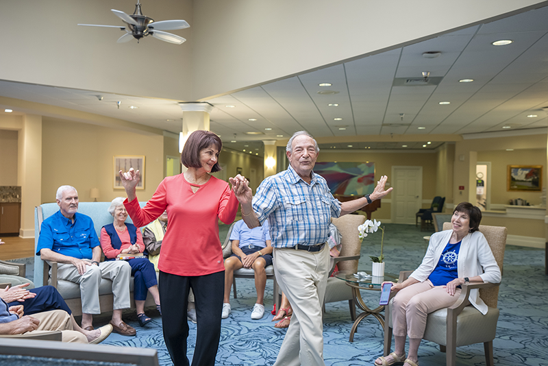 Two older adults dance together in the center of a common room while a group of seniors sit around them, watching and smiling.