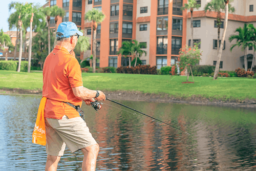 Man wearing an orange shirt and blue cap fishing by a pond, with apartment buildings and palm trees in the background.
