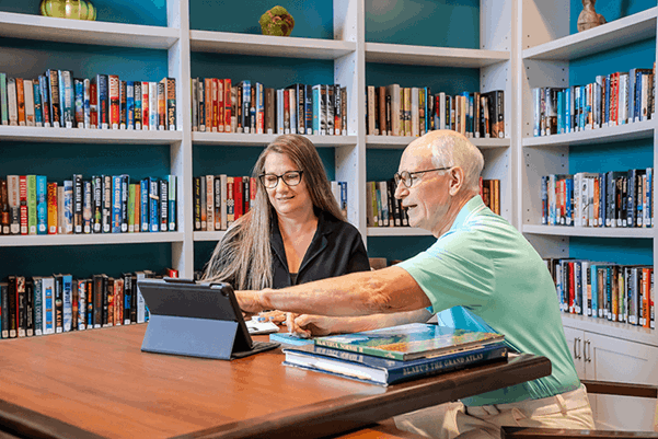 Two adults sit at a table in a library, looking at a tablet together. Bookshelves filled with books line the background.
