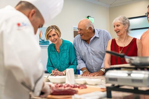 A chef prepares food while three adults and one other person watch from across a counter, smiling and engaged in the demonstration.