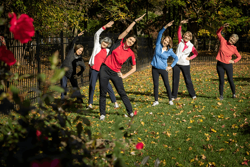 Six women stand on grass outdoors, stretching with arms overhead and leaning to one side. Trees with autumn leaves and a rose bush are visible in the background.