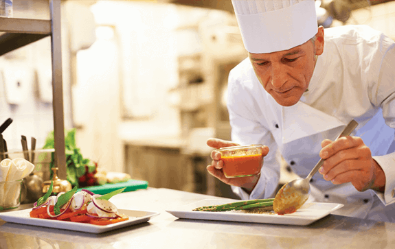 Chef in a white uniform and hat carefully plating food, pouring sauce onto asparagus on a white plate in a professional kitchen.
