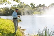 An older man wearing a cap and glasses stands on a grassy bank fishing in a calm lake on a sunny day.