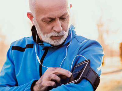 Older man in a blue jacket and earphones adjusts a smartphone strapped to his arm while outdoors.