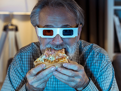 Older man in a plaid shirt wears 3D glasses and eats pizza indoors, appearing to watch something in front of him.