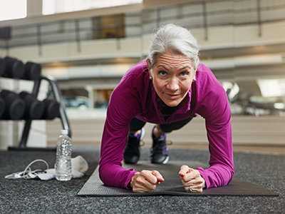 An older woman in athletic wear holds a plank position on a gym mat, with a water bottle and headphones nearby.