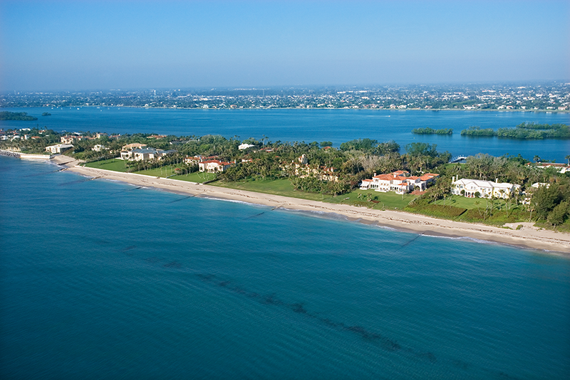 Aerial view of upscale beachfront homes along a sandy shoreline with calm blue water, lush greenery, and a distant cityscape under a clear sky.
