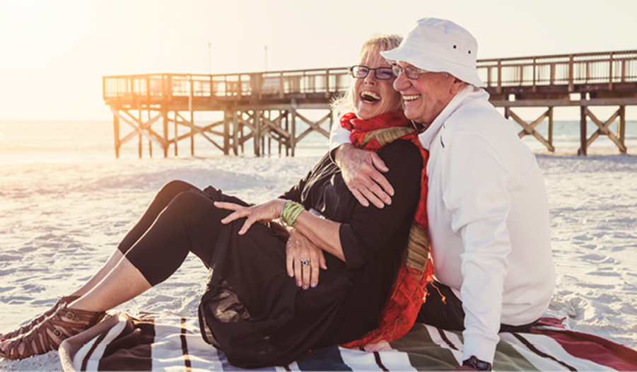 Elderly couple sitting on a beach blanket, smiling and embracing, with a pier and ocean in the background on a sunny day.