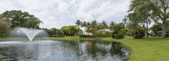 A small pond with a fountain is surrounded by grass, palm trees, and other greenery under a partly cloudy sky.