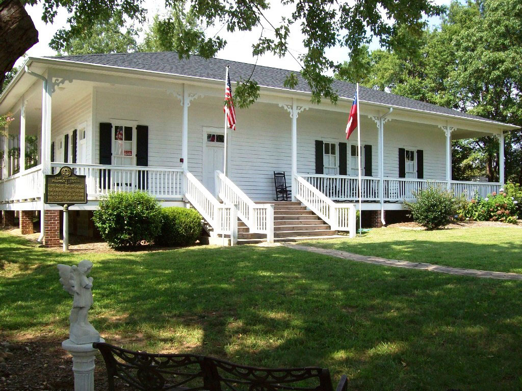 White historic house with a large front porch, two flags, and black shutters, surrounded by trees and a lawn with a small angel statue in the foreground.