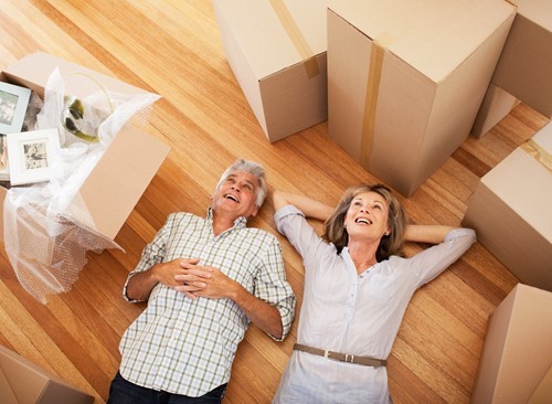 Two older adults lying on a wooden floor, smiling and surrounded by packed moving boxes and household items.