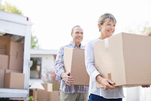 Two adults carrying cardboard boxes outside, near a moving truck filled with more boxes and household items, during a move.