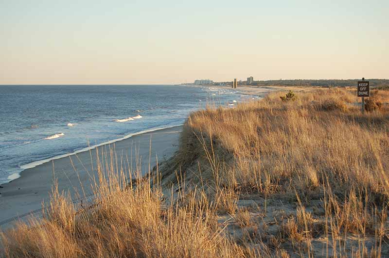 A sandy beach with gentle waves and tall grasses on dunes at sunset; a “Keep Off Dune” sign is visible on the right, and buildings appear in the distant background.