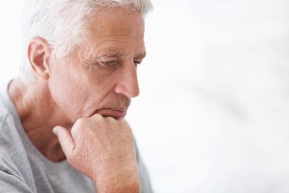 Older man with gray hair and a neutral expression rests his chin on his hand while looking downward. He is wearing a light gray shirt.