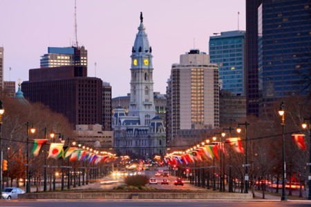 Philadelphia City Hall stands in the center, surrounded by tall buildings, with international flags lining the street and cars driving below during dusk.