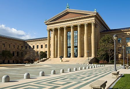 Large neoclassical building with tall columns, wide steps, and a patterned plaza in front under a clear blue sky.