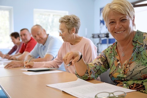 Five adults sit at a table in a classroom, writing on papers. The woman closest to the camera smiles, while others are focused on their work.