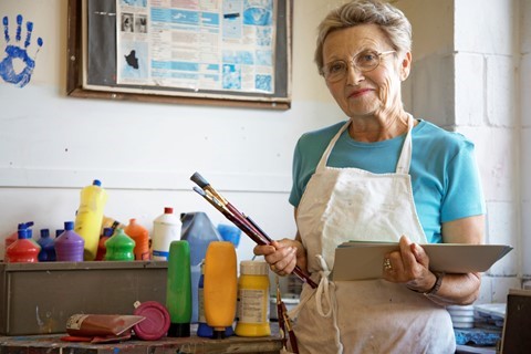 Older woman wearing glasses and an apron stands in an art studio, holding paintbrushes and a notebook, with colorful paint bottles on the table behind her.