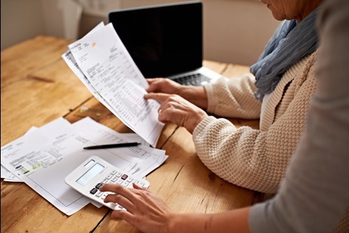 Two people review documents and use a calculator at a wooden table, with a laptop and more paperwork nearby.