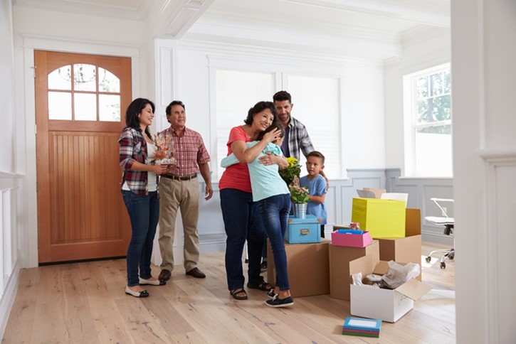 Six people stand in a bright room with moving boxes. Two women hug in the center while others watch and smile. A child holds flowers. The scene suggests moving into a new home.