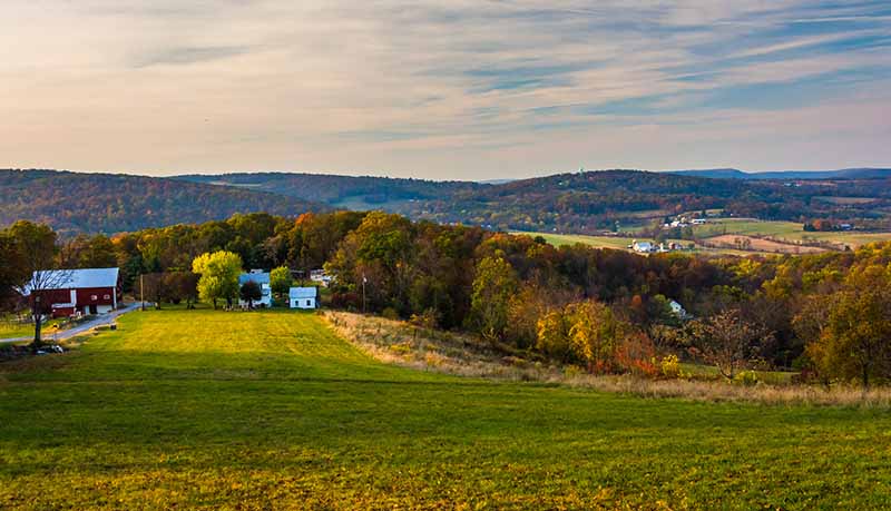 Rolling countryside with green fields, scattered farm buildings, trees with autumn foliage, and distant hills under a partly cloudy sky.