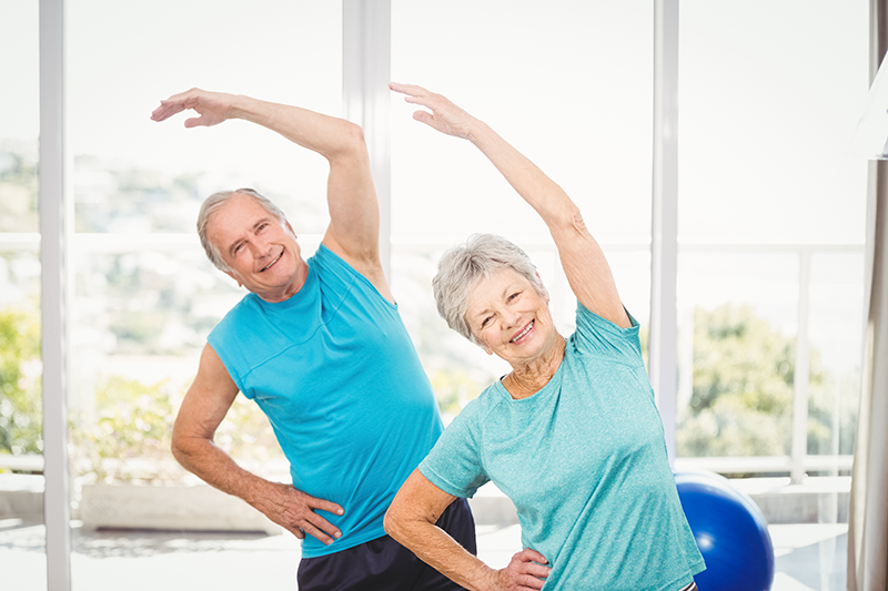 Two older adults in athletic wear stretch their arms overhead while exercising indoors near large windows.