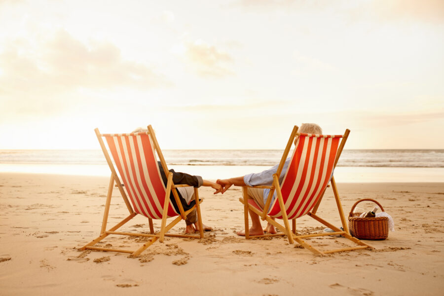Two people sit in striped deck chairs on a sandy beach at sunset, holding hands. A picnic basket is placed nearby.
