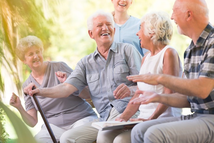 Four older adults and a younger person sit outdoors on a bench, smiling and laughing together in a sunny setting.