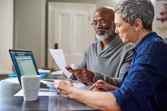 Two adults sit at a table, looking at documents and a laptop. One holds a paper, while the other points at the screen. A coffee cup and paperwork are on the table.