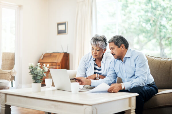 Older couple sitting on a couch looking at a laptop together, with papers and coffee cups on the table in a bright living room.