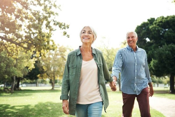 Two older adults walk hand in hand through a sunny park, smiling. Trees and green grass are visible in the background.