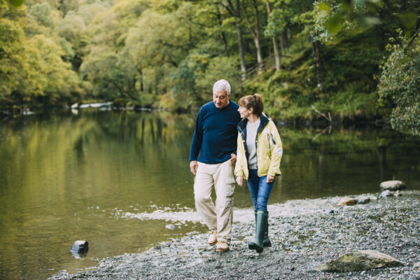 Two people walk together along a rocky riverbank in a forested area, with trees and calm water in the background.
