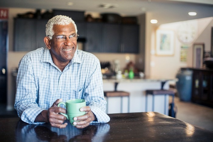 An older man with gray hair and glasses sits at a table holding a green mug, smiling, in a modern kitchen with dark cabinets and a blurred background.