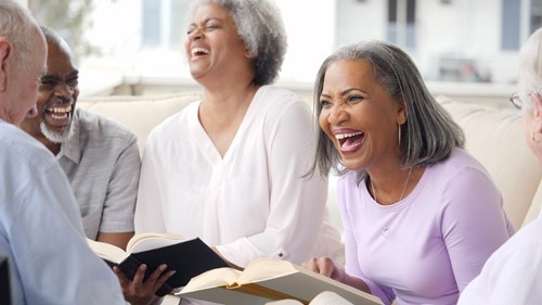 Four older adults sit closely together on a sofa, smiling and laughing while holding open books, suggesting they are enjoying a group reading or book club activity.