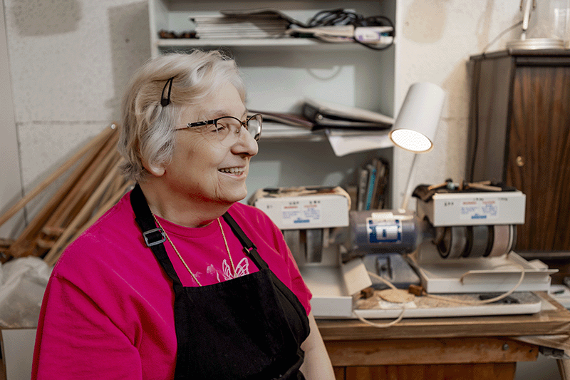 An older woman wearing glasses and a dark apron smiles while sitting in a workshop with two grinding machines and various tools behind her.