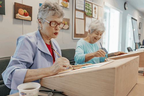 Two elderly women sit at tables, painting on canvases in a well-lit room with artwork displayed on the walls.