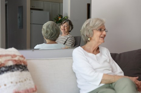 An older woman in glasses sits on a couch, with her reflection visible in a mirror behind her.